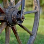 Close-up of an old, weathered wagon wheel made of wood with a dark iron hub, set against a blurred grassy background.