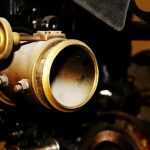 Close-up of a vintage projector lens with a large circular brass lens ring, an adjustment knob, and mounting bolts, set against a background of dark metal machinery.
