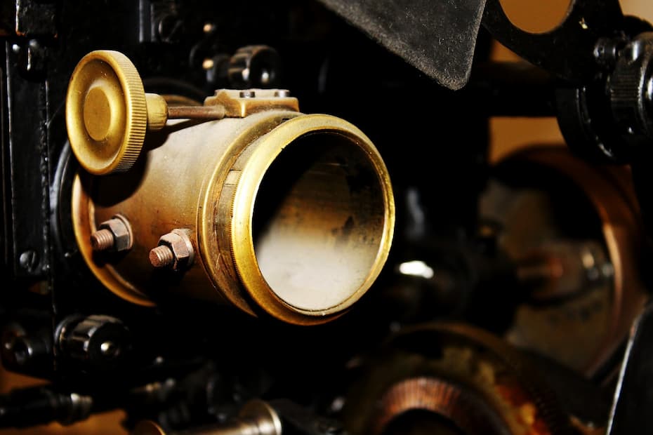 Close-up of a vintage projector lens with a large circular brass lens ring, an adjustment knob, and mounting bolts, set against a background of dark metal machinery.