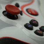 A close-up of a white and red video game controller, showing a red thumbstick and four buttons labeled A, B, X, and Y in blue, red, pink, and teal.