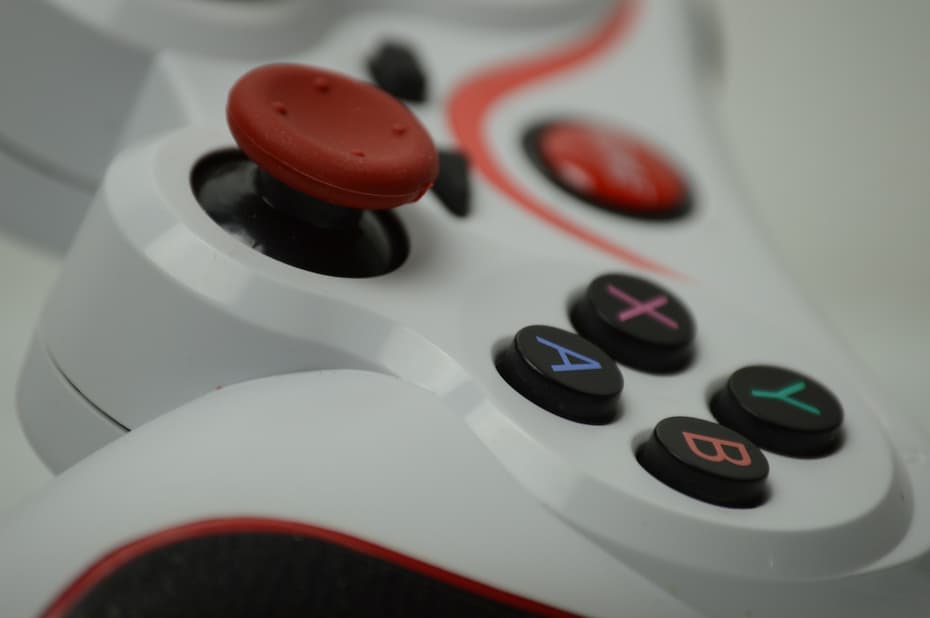 A close-up of a white and red video game controller, showing a red thumbstick and four buttons labeled A, B, X, and Y in blue, red, pink, and teal.