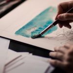 Close-up of hands painting a watercolor boat scene next to pencil sketches.