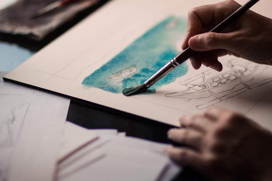 Close-up of hands painting a watercolor boat scene next to pencil sketches.